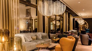 A modern living room with a large sofa, armchairs, and a coffee table. There are decorative lights and a chandelier. - Motel One Aachen (Aachen)