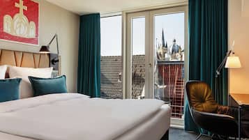 A hotel room with a large bed, a desk with a chair, a window with curtains, and a view of a cityscape.