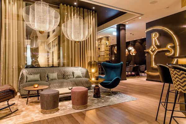 A modern hotel lobby with a sofa, round ottoman, and a blue chair. - Motel One Aachen (Aachen)