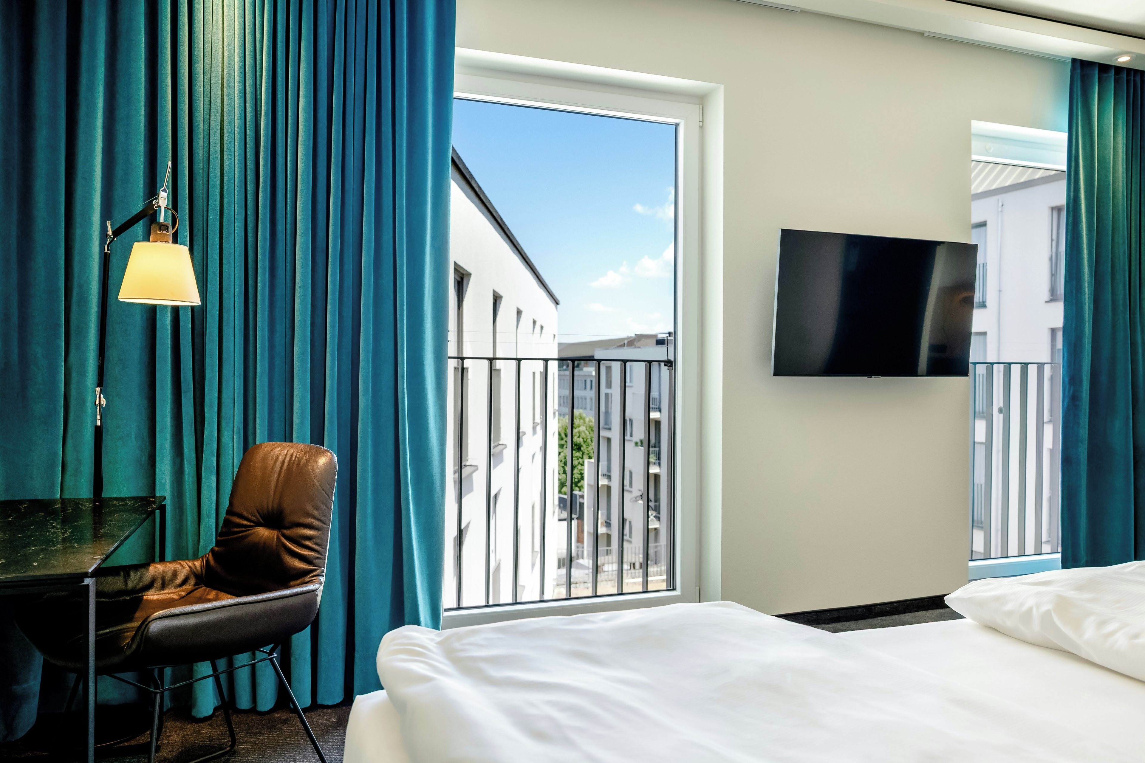 A hotel room with a bed, a desk, a chair, a TV, and a view of buildings through the window.