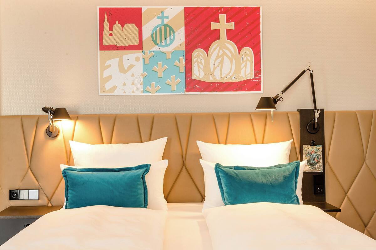 Une chambre d’hôtel avec une tête de lit capitonnée, deux lits avec des oreillers blancs et turquoise, une œuvre d’art accrochée au mur et une table de chevet avec une lampe.