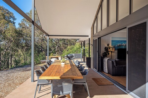 An outdoor dining area with a long wooden table and chairs, a barbecue grill, and a view of the surrounding trees.