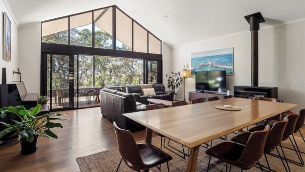 A modern living space with a large dining table, comfortable seating, a fireplace, and large windows offering a view of the outdoors. - Gum Nuts - Nestled amongst the leafy trees, Australian styled simple & relaxing (Yallingup)