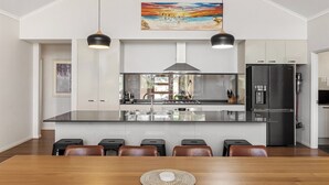 A modern kitchen with a central island, stainless steel appliances, pendant lighting, and a dining area with a wooden table and chairs. - Gum Nuts - Nestled amongst the leafy trees, Australian styled simple & relaxing (Yallingup)