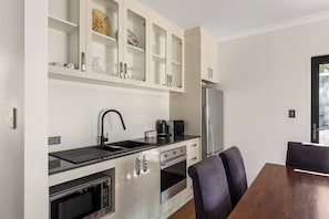 Een kleine keuken met een spoelbak, fornuis en koelkast, een eettafel met stoelen, en een kastje met decoratieve spullen.