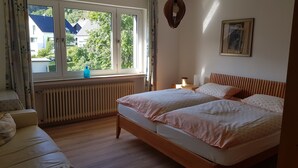 Un dormitorio con dos camas, una ventana con cortinas, un radiador, una mesita de noche con lámpara, y un cuadro en la pared.