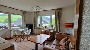 Una sala de estar moderna con una mesa de centro de madera, un sofá de cuero y un televisor. Amplias ventanas ofrecen vistas al campo.