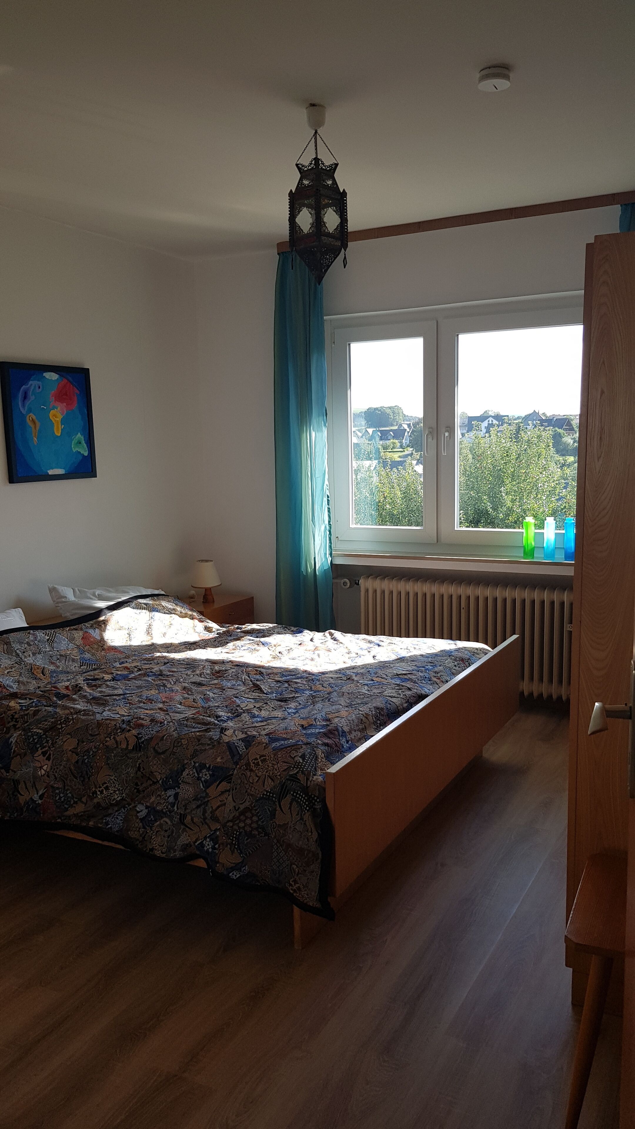 A bedroom with a large bed, a window with a view, a radiator, and a ceiling light fixture.