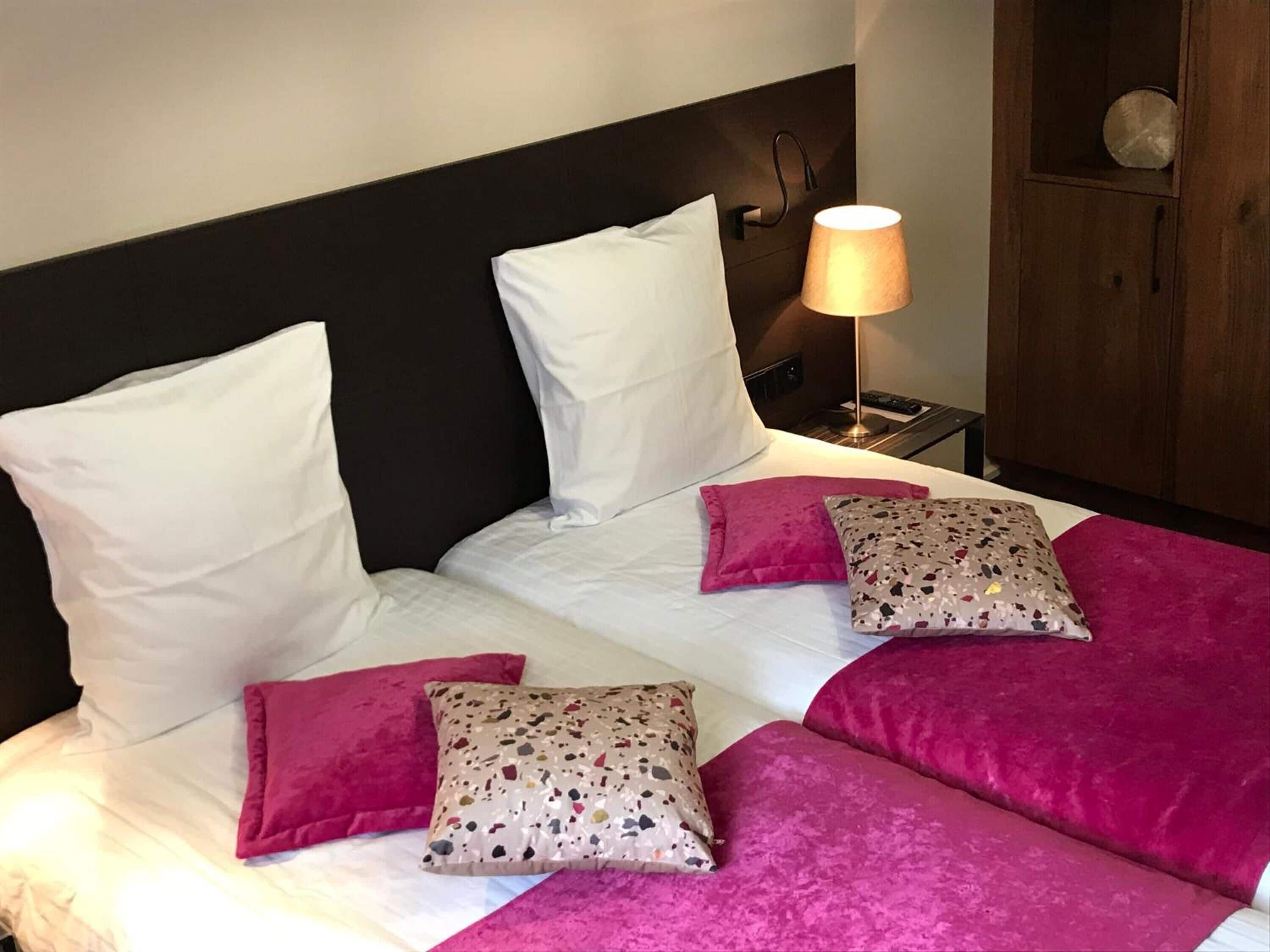 Cama bem arrumada com roupa de cama branca e rosa, cabeceira e abajur.