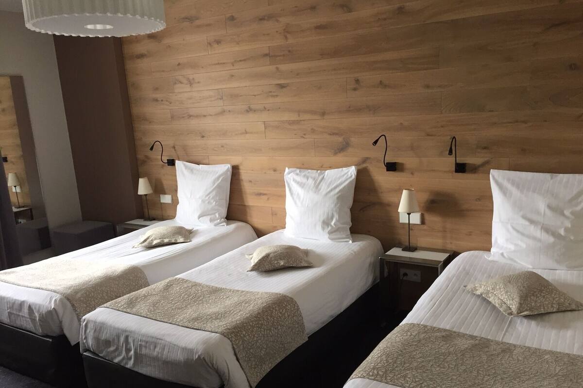 A hotel room with three single beds, each with a white pillow and a beige throw blanket. The room has wooden paneling on the walls and a modern, minimalist design.
