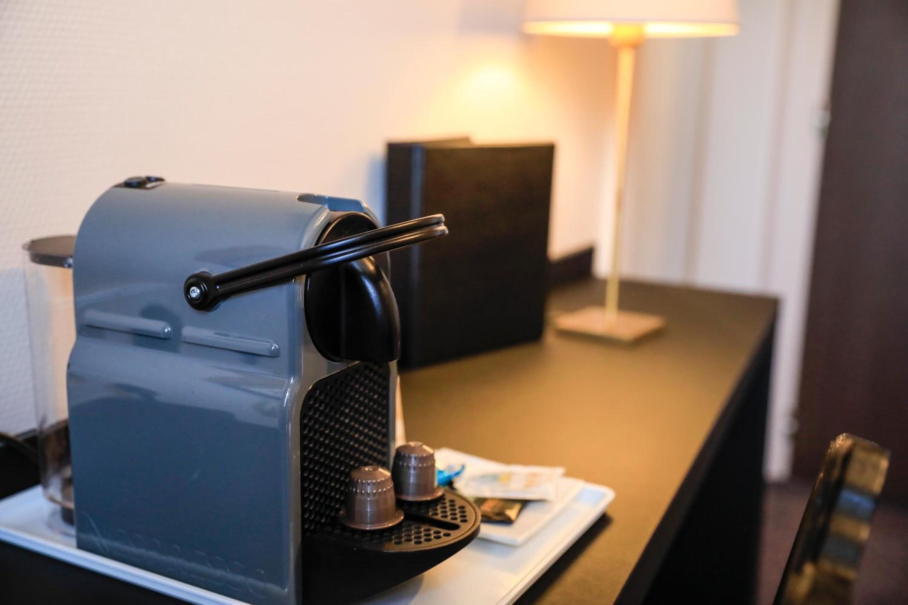 A compact, modern coffee machine on a white tray, with a small table and a lamp in the background.