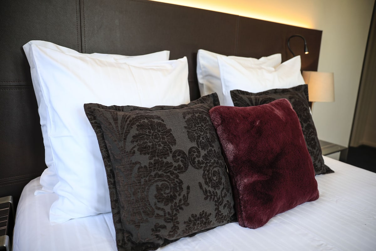 A neatly made bed with white and patterned pillows, a red throw pillow, and a bedside lamp.