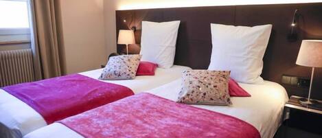 Habitación de hotel con dos camas, cada una con cabecero blanco y colchas rojas. Hay dos lámparas de noche y una ventana con cortinas.