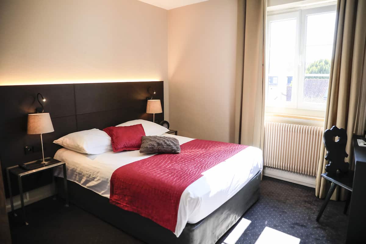A hotel room with a large bed, two bedside tables with lamps, a view of the outside through a window, and a chair near the window.
