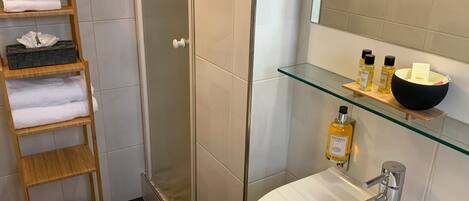 A modern bathroom with a glass shower enclosure, a wooden towel rack, a glass shelf with toiletries, and a white sink.