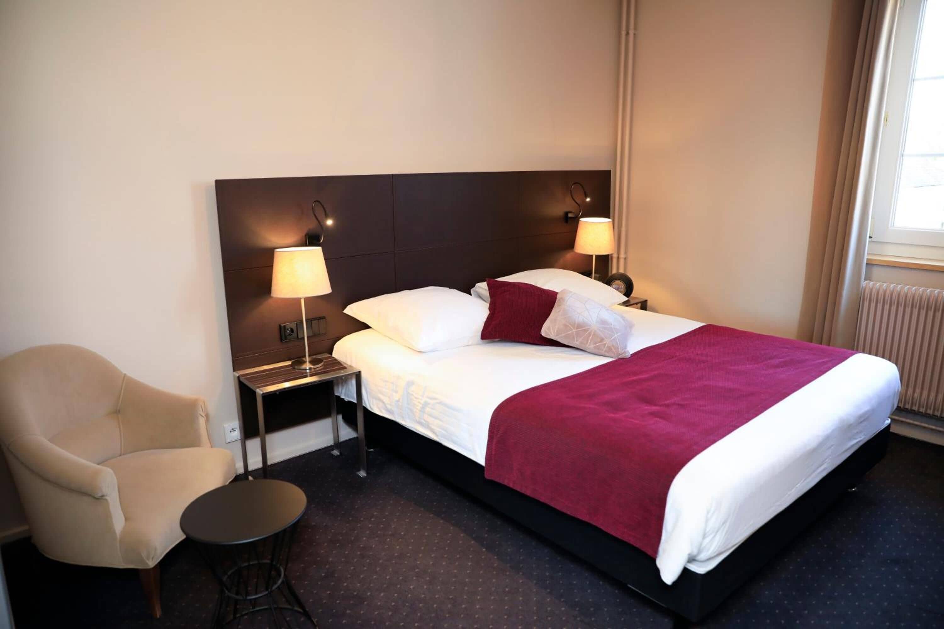 Ein Hotelzimmer mit einem großen Bett, zwei Nachttischlampen, einem Sessel und einem kleinen runden Tisch.