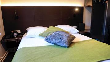Una habitación de hotel con cama, ropa de cama verde y blanca, una mesita de noche con teléfono y una lámpara fijada a la pared.