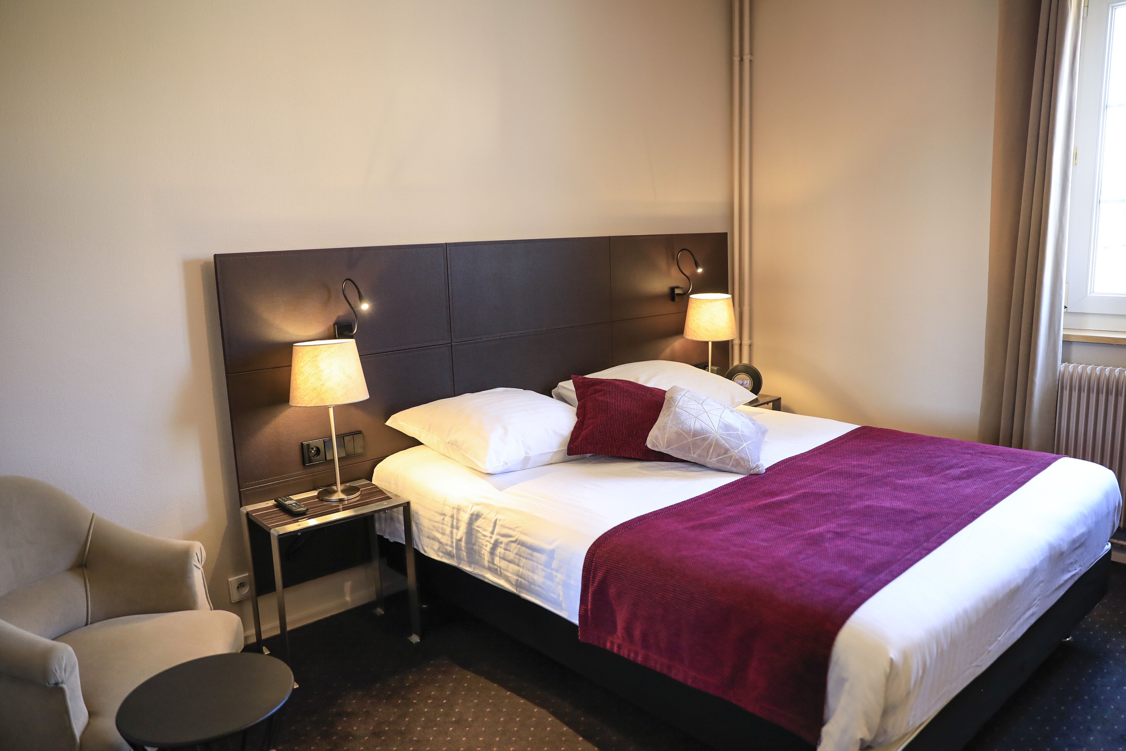 A hotel room with a bed, two bedside lamps, a chair, a small table, and a window with curtains.