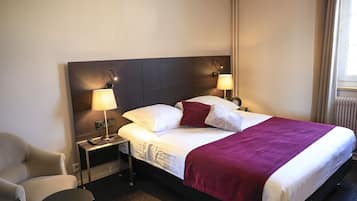 A hotel room with a bed, two bedside lamps, a chair, a small table, and a window with curtains.