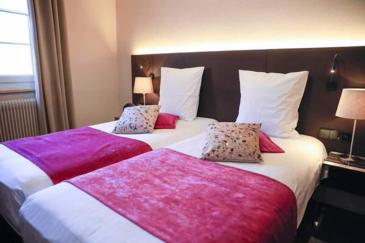 A hotel room with two beds, each with a headboard, bedside lamps, and a window with curtains.