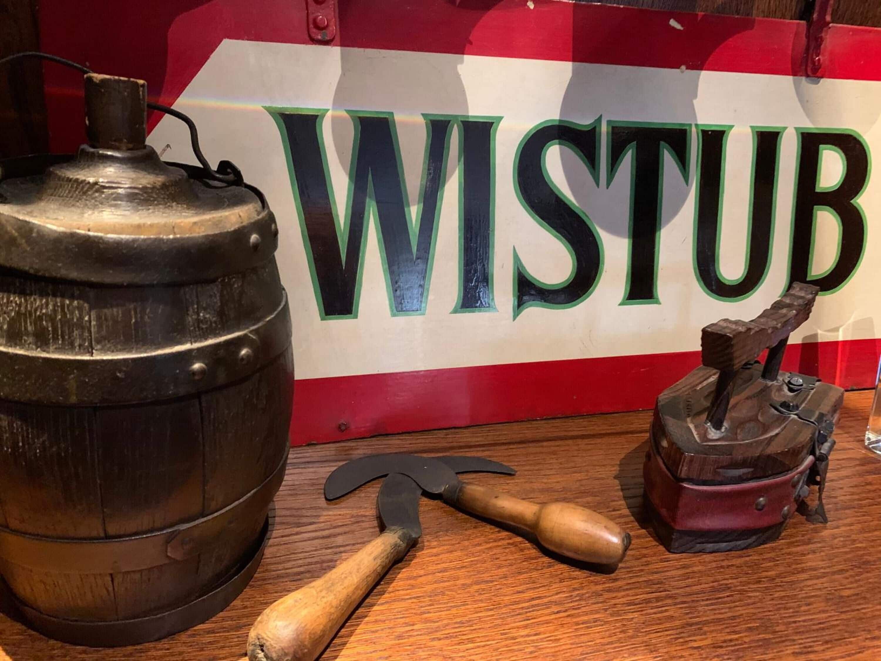 Ein Holfass, ein Handwerkzeug und eine alte Presse vor einem Hintergrund mit der Aufschrift 'WISTUB'.