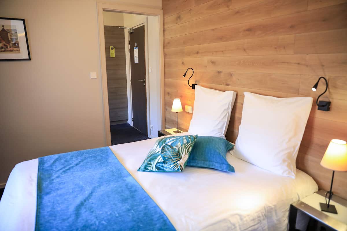 A hotel room with a wooden headboard, a bed with white and blue bedding, two bedside lamps, and a door leading to another room.