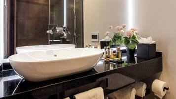 A modern bathroom with a large white vessel sink, a black countertop, a glass shower enclosure, and a potted plant with flowers.