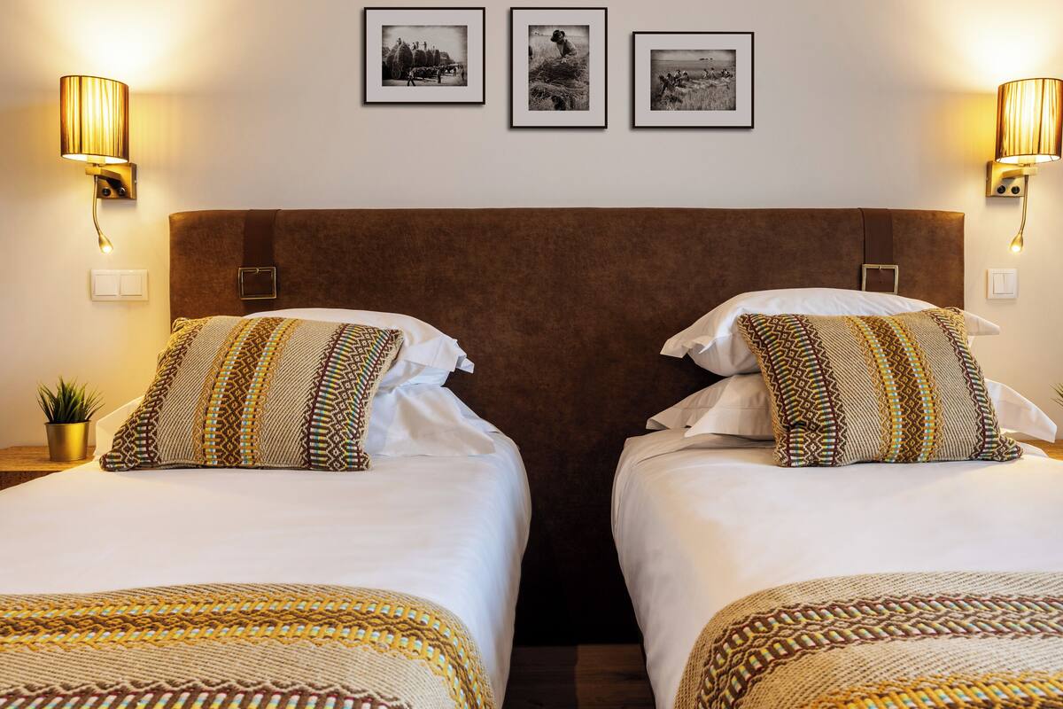 A hotel room with two beds, each with patterned pillows and a brown headboard. There are wall-mounted lamps and framed pictures above the beds.