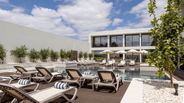 Zona de piscina con sillones de descanso y sombrillas, un edificio moderno al fondo y cielo despejado.