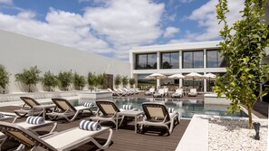 Zona de piscina con sillones de descanso y sombrillas, un edificio moderno al fondo y cielo despejado.