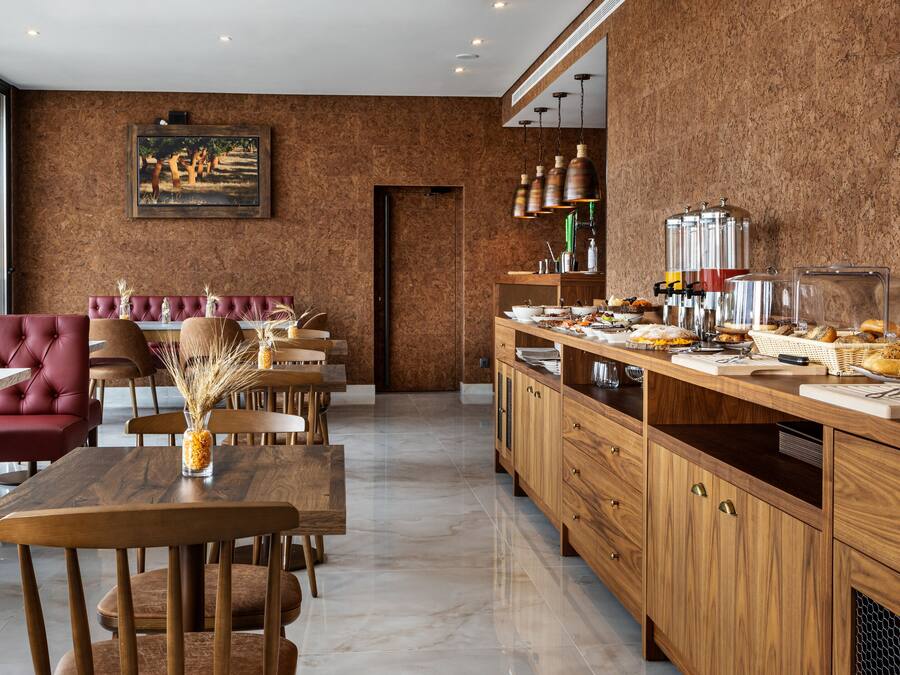 A modern dining area with a red upholstered bench, wooden tables, and chairs. A buffet counter with various food items is visible.