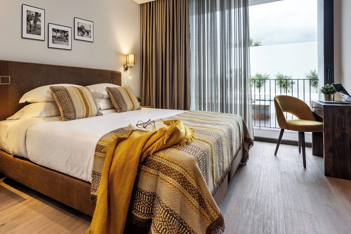 A modern hotel room with a large bed, a desk with a laptop, and a balcony with a chair.