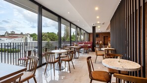 Un restaurante moderno con amplios ventanales, mesas de madera y vistas a edificios y árboles.