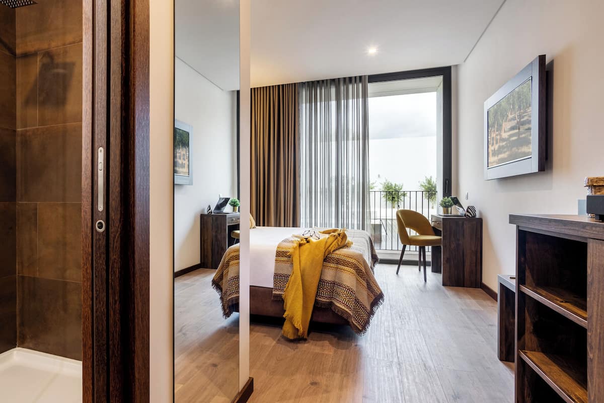 A modern hotel room with a glass shower enclosure, a bed with a patterned blanket, a desk with a laptop, and a view of the outdoors through large windows.