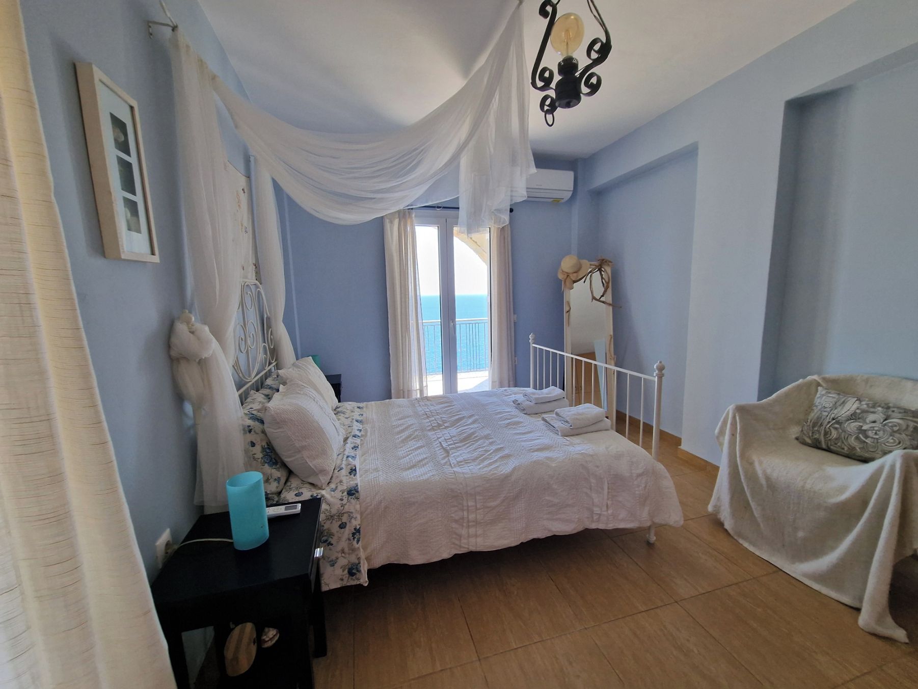 A bedroom with a bed, a nightstand, a chair, a mirror, and a view of the sea through the window.