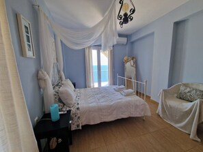 A bedroom with a bed, a nightstand, a chair, a mirror, and a view of the sea through the window. - Villa Aegean Balcony (Korasida)
