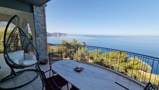 Ein Balkon mit einer Hängematte, einem Tisch und Stühlen. Blick auf ein Gewässer und die Ferne.