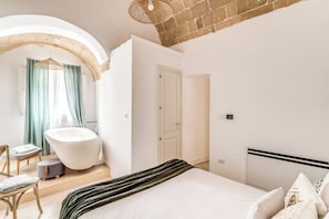 Una camera da letto moderna con un letto grande, un angolo relax con una poltrona e poggiapiedi, una vasca da bagno e una porta che dà su un'altra stanza.