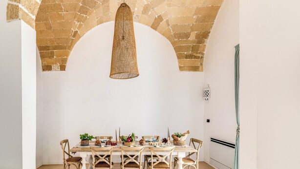 A dining area with a stone arch ceiling, a woven pendant light, and a white table set with various dishes and drinks.