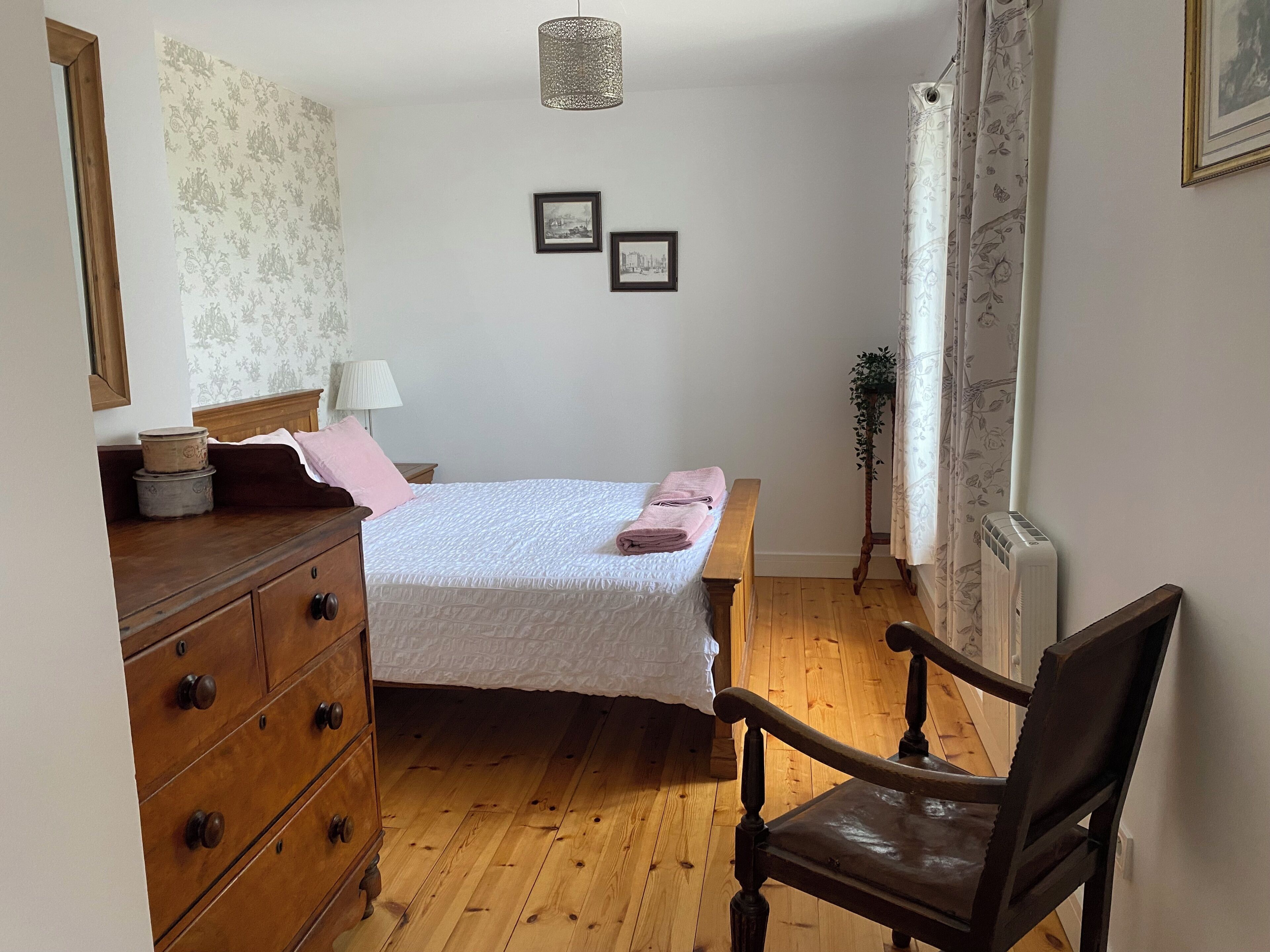 Une chambre avec une commode en bois, un lit recouvert d’une courtepointe blanche, une chaise, une lampe et deux tableaux encadrés accrochés au mur.
