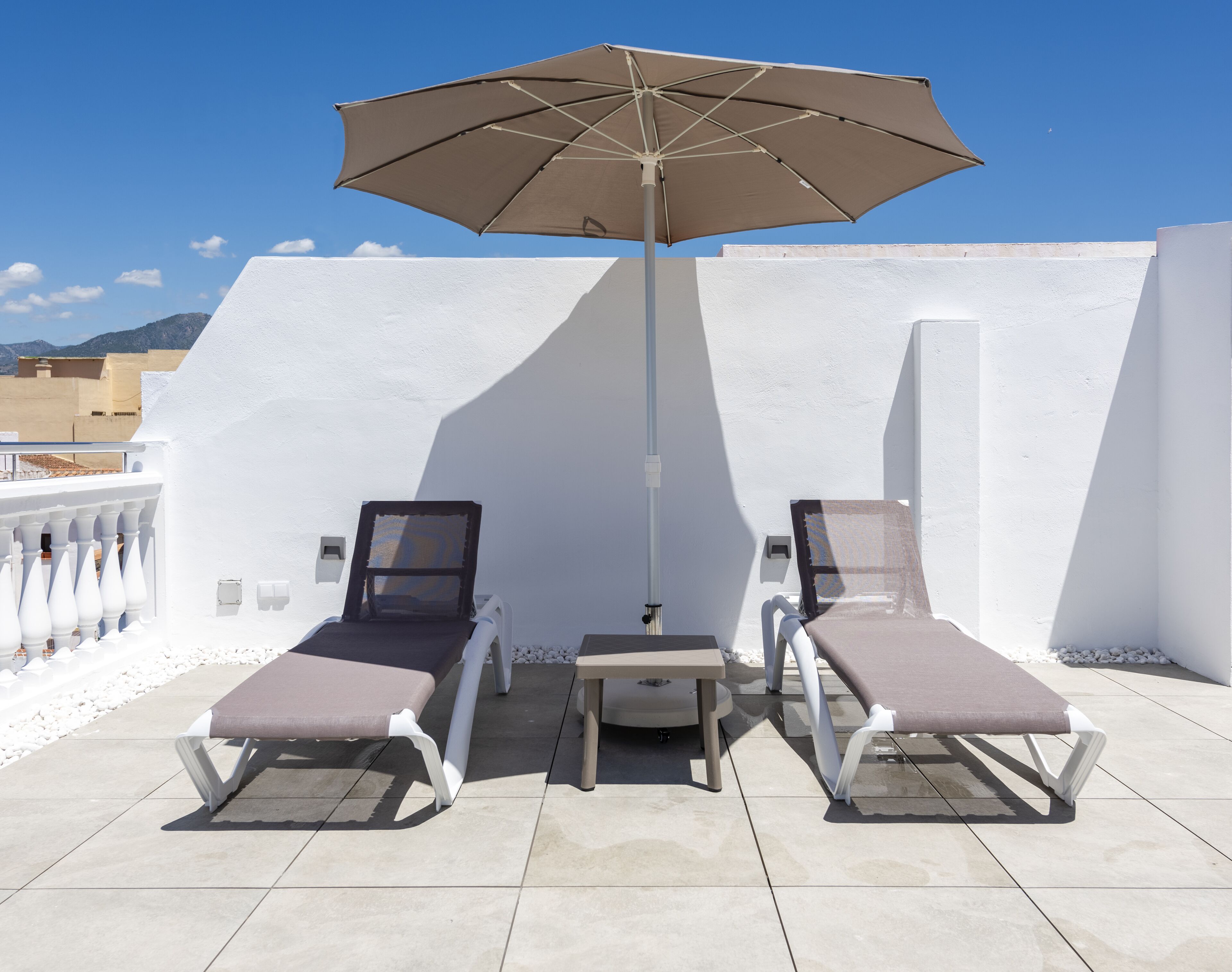 Two sun loungers with a table and an umbrella on a tiled patio with white walls and a clear sky.