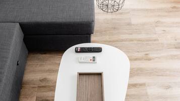 Una sala moderna con un sofá gris, una mesa de centro blanca con un marco de fotos, un control remoto y una planta pequeña sobre una mesita redonda.