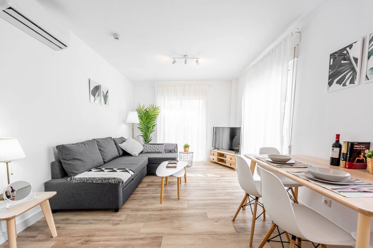Een moderne woonkamer met een grijze bank, een eettafel met stoelen, een televisie en een plant.
