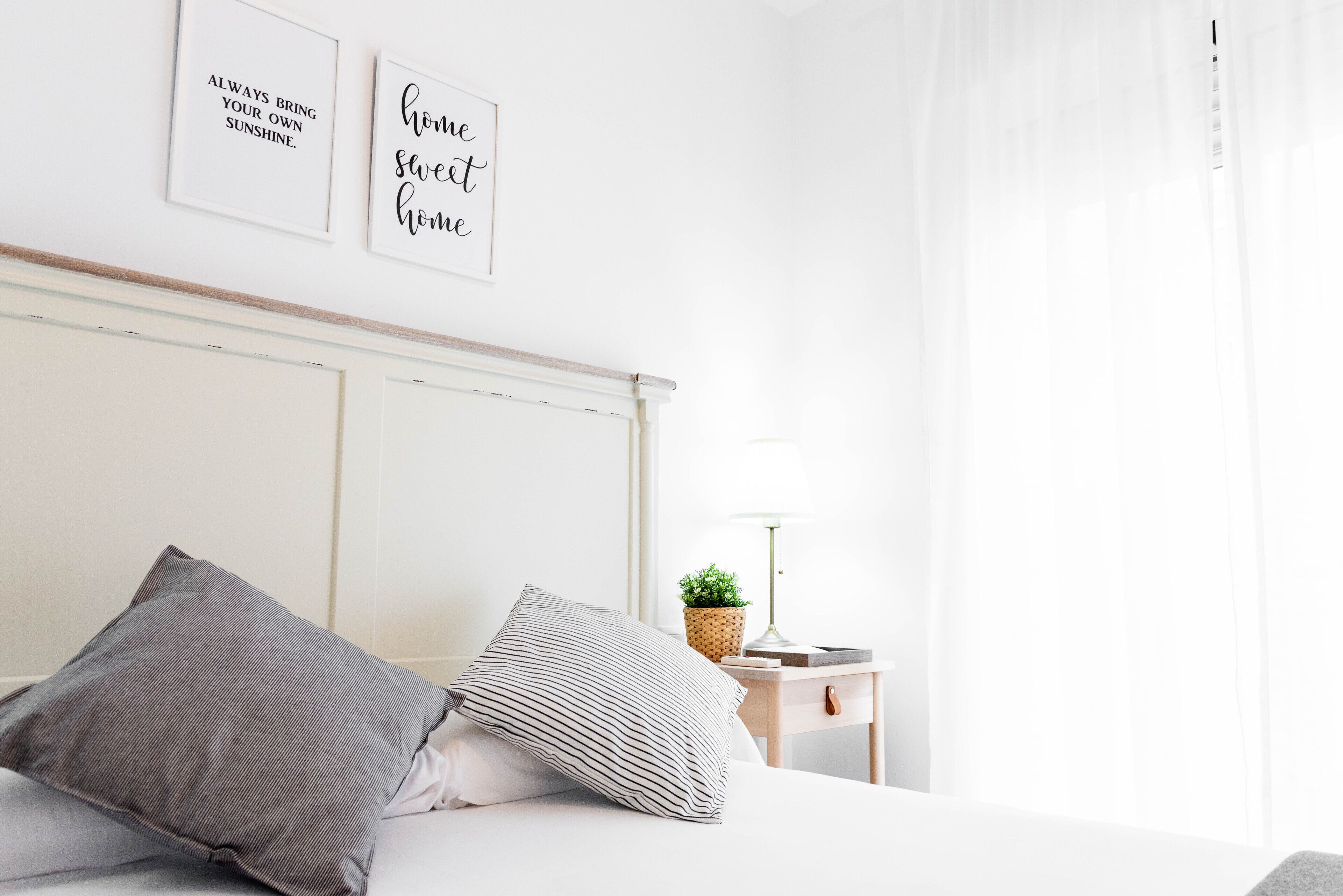 Una cama bien hecha con almohadas a rayas, una mesita de noche con una lámpara y una planta, y dos cuadros con citas motivacionales.