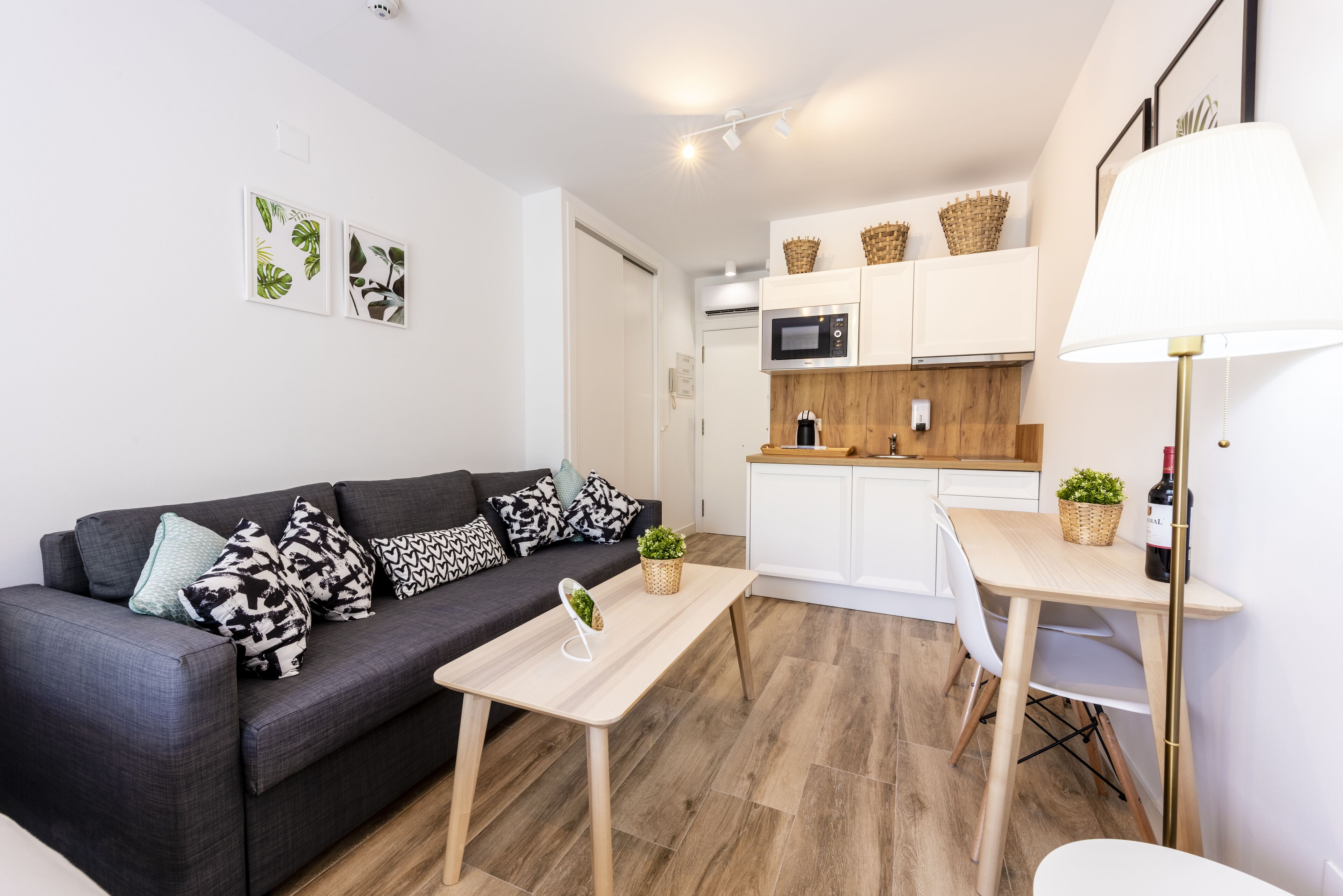 Un apartamento moderno con un sofá gris oscuro, una mesita de madera, una zona de cocina con armarios blancos y una mesa de comedor.