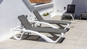 Eine Dachterrasse mit zwei Liegestühlen und einem kleinen Tisch, umgeben von weißen Wänden und einem gefliesten Boden.