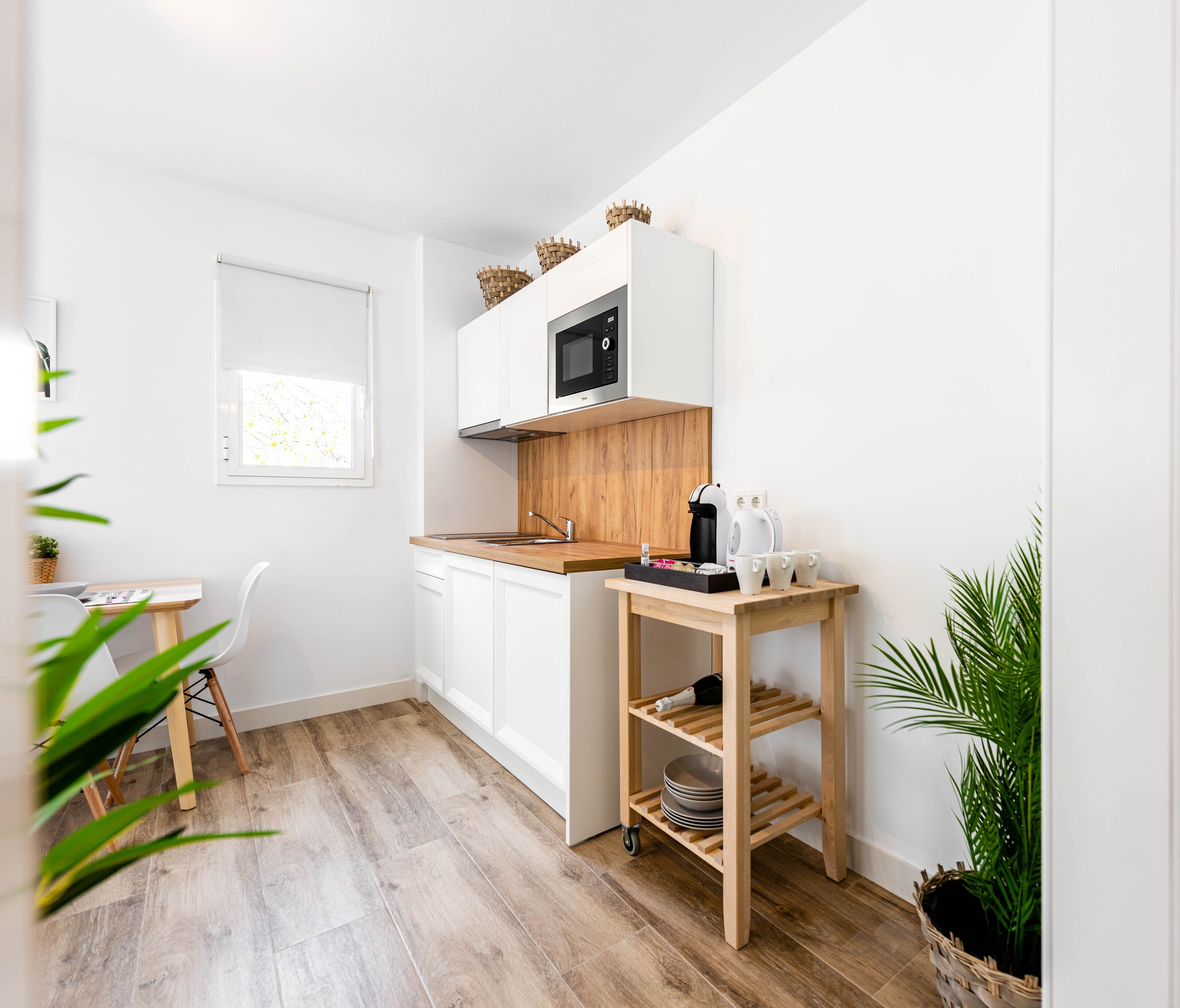 Una cocina compacta con muebles blancos, un microondas, encimera de madera y una mesita con tazas y cafetera.