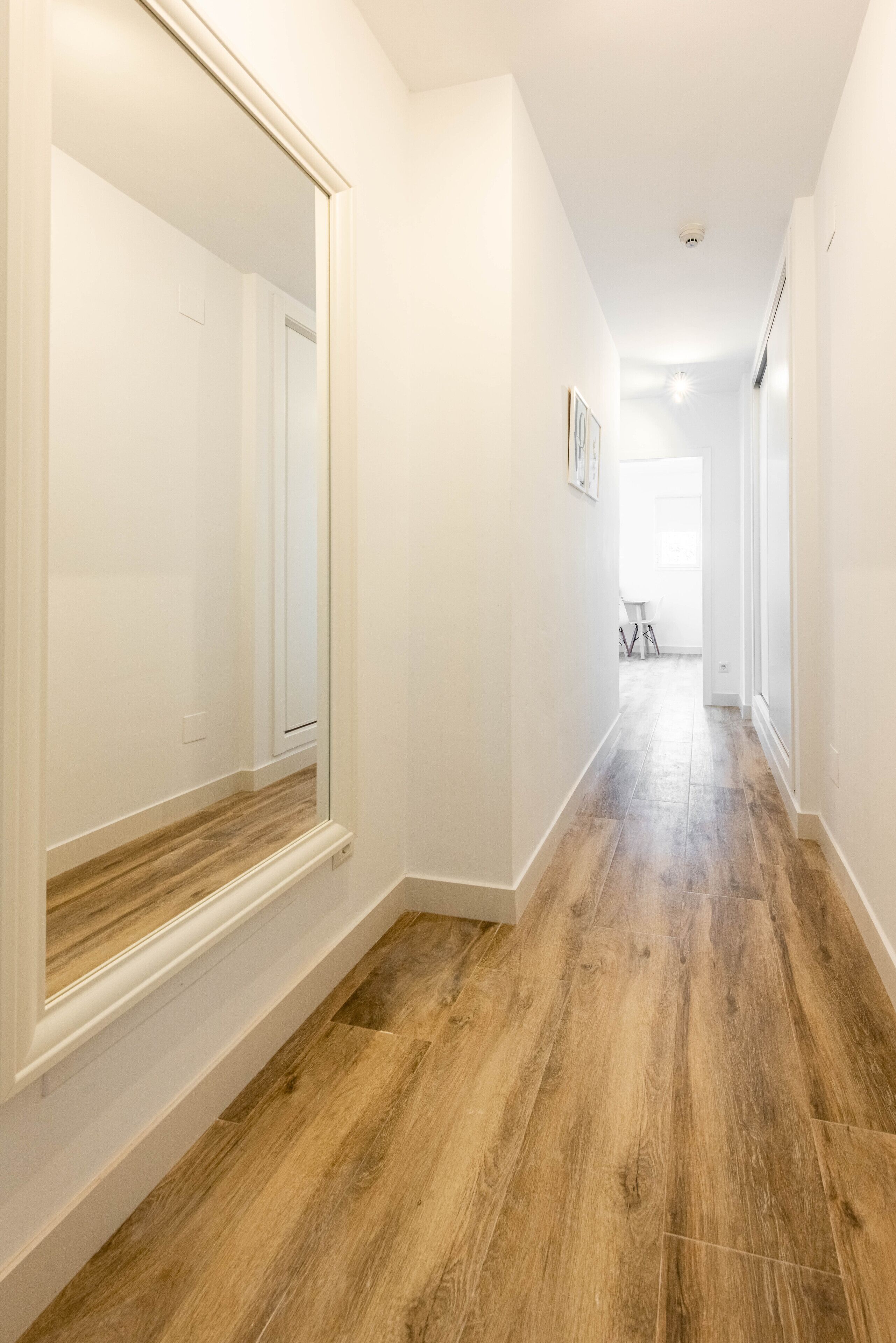 Un couloir doté d’un plancher en bois, de murs blancs et d’un grand miroir sur le côté gauche.