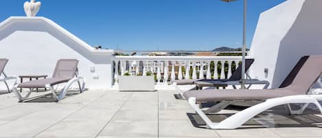 A rooftop terrace with sun loungers, a large umbrella, and a white railing.