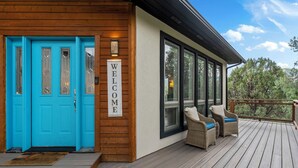 A wooden exterior with a bright blue door, a 'WELCOME' sign, and wicker furniture on a deck with a view of trees.